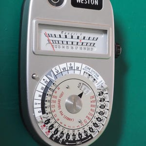 May include: A vintage Weston Ranger 9 light meter with a silver body. The meter features a dial with numbers and markings for light measurement, including ASA and time settings. The word "WESTON" is printed on the top.