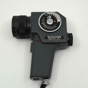 May include: A gray Spiratone SP-1 light meter with a black lens and a circular dial with red markings. The dial has numbers from 2 to 16 and the words "ASA" and "DIN".