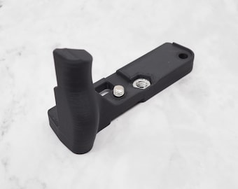 May include: A black camera grip with a textured handle and a mounting plate. The grip has a curved design for comfortable handling and includes a screw and a threaded insert for secure attachment to a camera.
