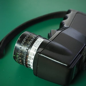 May include: A black Zone VI Studios Inc. light meter with a black strap. The light meter has a dial with numbers and a zone scale.