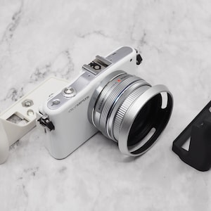 May include: A white Olympus camera with a silver lens and a black lens hood. The camera is attached to a white accessory and a black accessory.