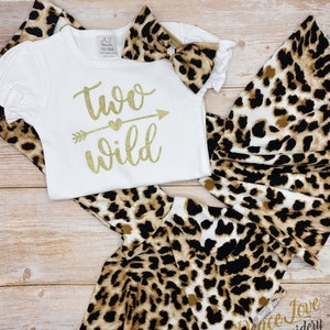 May include: A white onesie with gold glitter text that reads "two wild" with an arrow and heart. The onesie is paired with a leopard print headband and matching leggings.
