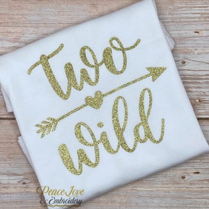 May include: White t-shirt with gold glitter lettering that says "Two Wild" with an arrow and heart design. The text is in a cursive font.
