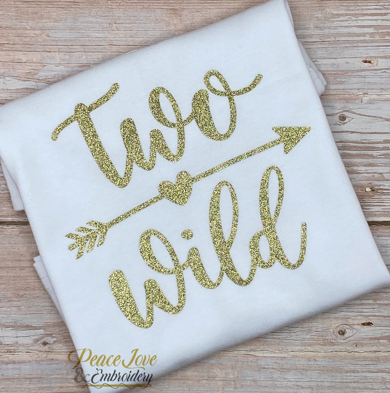 May include: White t-shirt with gold glitter lettering that says "Two Wild" with an arrow and heart design. The text is in a cursive font.