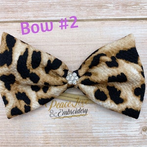May include: A brown and black leopard print bow with a clear rhinestone in the center. The bow is attached to a nude elastic headband. The text "Bow #2" is written in pink above the bow.
