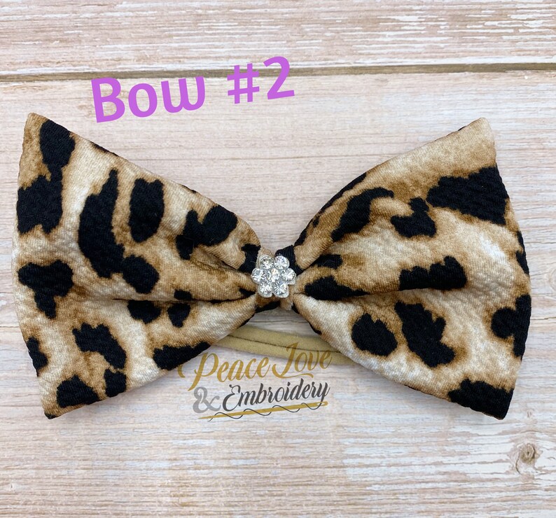 May include: A brown and black leopard print bow with a clear rhinestone in the center. The bow is attached to a nude elastic headband. The text "Bow #2" is written in pink above the bow.