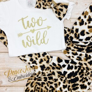May include: A white onesie with gold glitter text that says "two wild" with an arrow and heart. The onesie is paired with a leopard print headband and matching leggings.