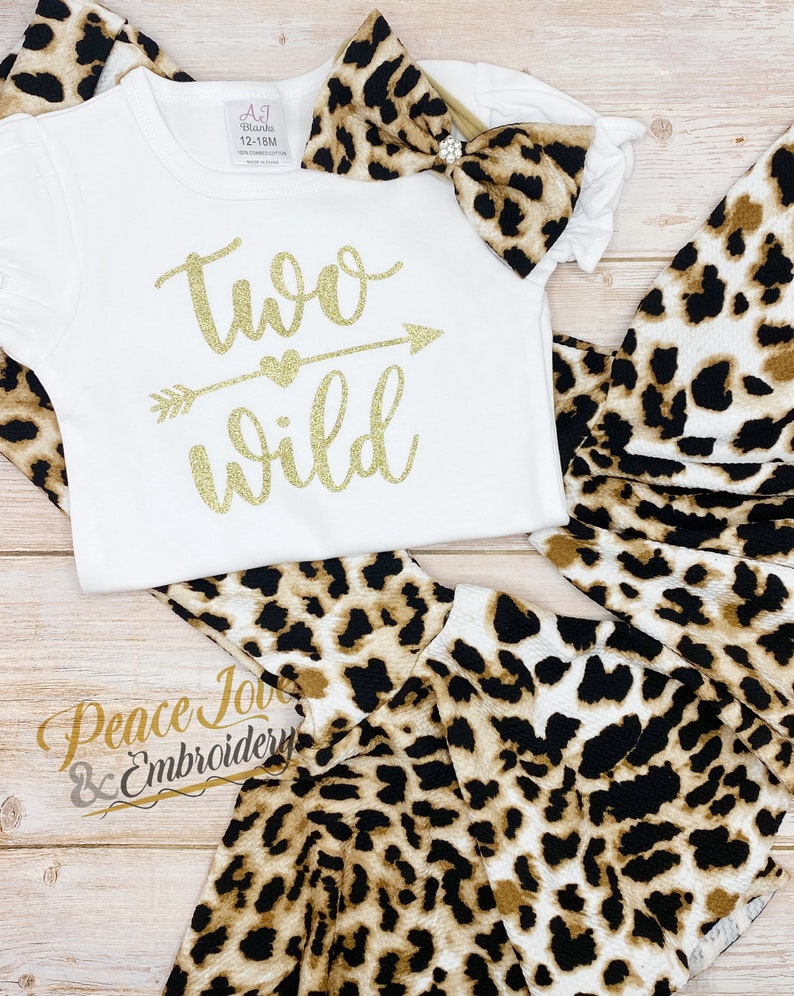 May include: A white onesie with gold glitter text that says "two wild" with an arrow and heart. The onesie is paired with a leopard print headband and matching leggings.
