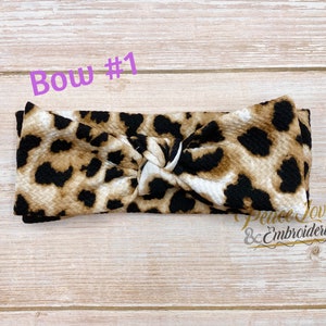 May include: A brown, black, and white leopard print knotted headband. The headband is made of a soft, stretchy fabric and is perfect for adding a touch of style to any outfit. "Bow #1" is written in purple text above the headband.
