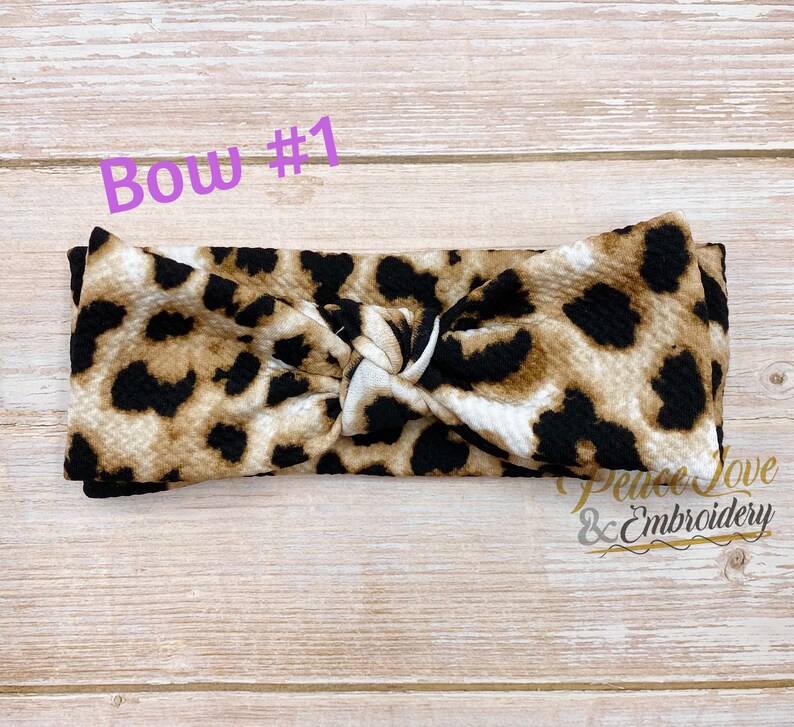 May include: A brown, black, and white leopard print knotted headband. The headband is made of a soft, stretchy fabric and is perfect for adding a touch of style to any outfit. "Bow #1" is written in purple text above the headband.