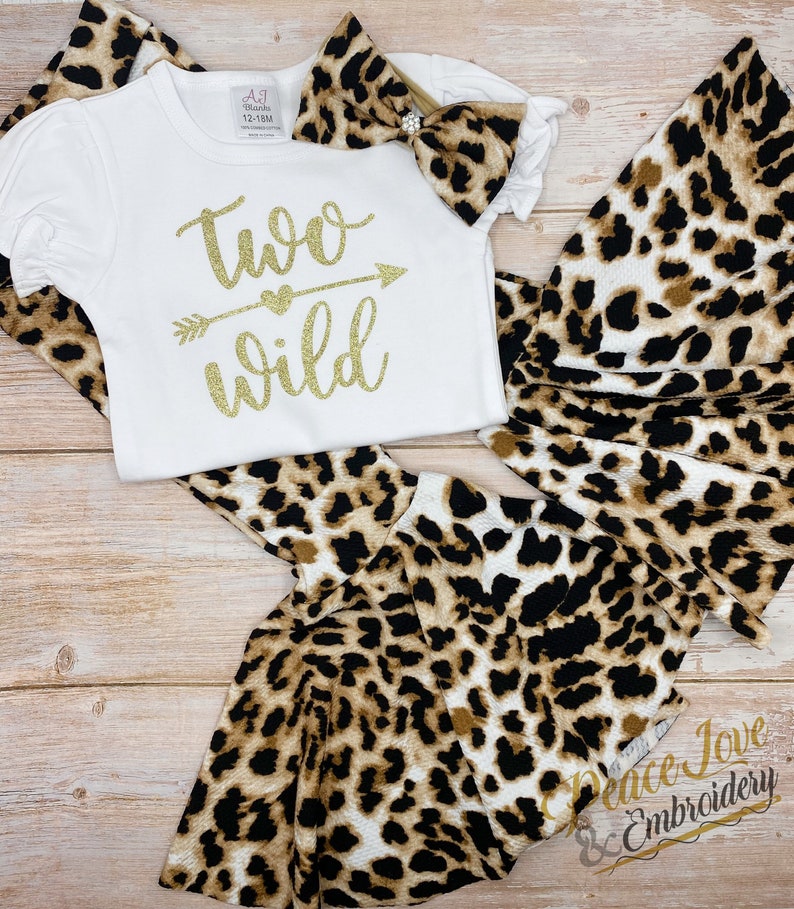 May include: A white onesie with gold glitter text that reads "two wild" with an arrow and heart. The onesie is paired with a leopard print headband and matching leggings.