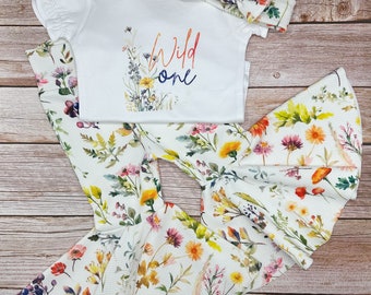 Puede incluir: Un body blanco con las palabras "Wild One" en letras de colores y un diseño floral. El body se combina con una diadema floral y leggings florales a juego.