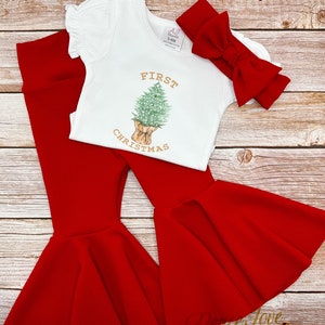May include: A white onesie with the text "FIRST CHRISTMAS" and a drawing of a Christmas tree in a brown paper bag. The onesie is paired with red bell bottom pants and a red bow headband.