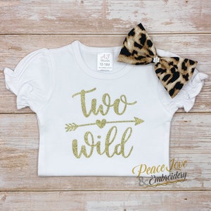 May include: White toddler shirt with gold glitter text that says "two wild" with an arrow and heart. A leopard print bow with a rhinestone is on top of the shirt.