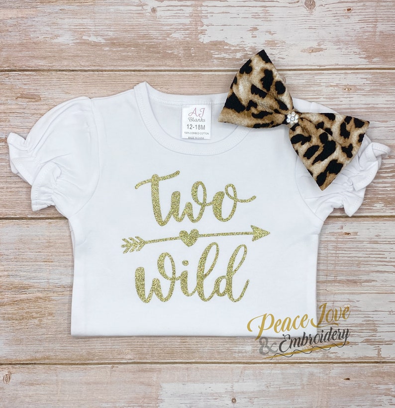 May include: White toddler shirt with gold glitter text that says "two wild" with an arrow and heart. A leopard print bow with a rhinestone is on top of the shirt.