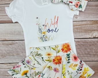 Puede incluir: Un body blanco con el texto "Wild One" y diseños florales. También se incluye una diadema floral a juego y una falda blanca con estampado floral.