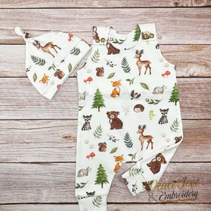 May include: A white baby onesie with a woodland animal print featuring deer, foxes, raccoons, squirrels, bears, and trees. A matching white hat with the same print is included.