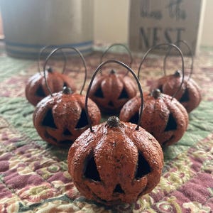 May include: A collection of small, orange pumpkin-shaped lanterns with black cut-out faces and wire handles. The lanterns are arranged on a patterned fabric, suggesting a fall or Halloween theme. A decorative sign with the text "Bless the NEST" is in the background.