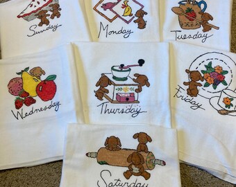 May include: A set of seven white kitchen towels, each embroidered with a different day of the week and a corresponding whimsical design. The designs include baked goods, kitchen utensils, and fruit, all with a cute dog theme.