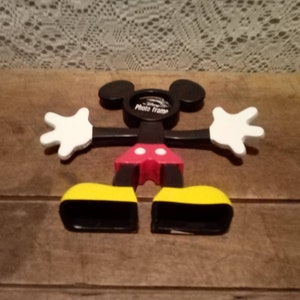 May include: Black and white Mickey Mouse photo frame with red shorts and yellow shoes. The frame is shaped like Mickey Mouse's head and has a space for a photo in the middle. The frame is made of plastic and is about 10 inches tall.