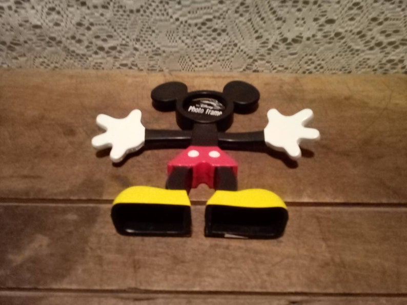 May include: Black and white Mickey Mouse photo frame with red shorts and yellow shoes. The frame is shaped like Mickey Mouse's head and has a space for a photo in the middle. The frame is made of plastic and is about 10 inches tall.