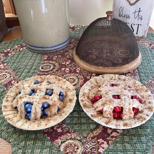 May include: Two decorative miniature pies, one with blue berries and the other with red berries, sit on a green and burgundy patterned quilt. A beige ceramic jug with blue stripes and a mesh food cover are also present.