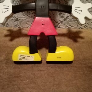 May include: A black and red articulated Mickey Mouse figurine with yellow shoes. The shoes have text on the bottom that reads "The Disney Store, Inc. Glendale, CA 91203-2671, USA" and "039".