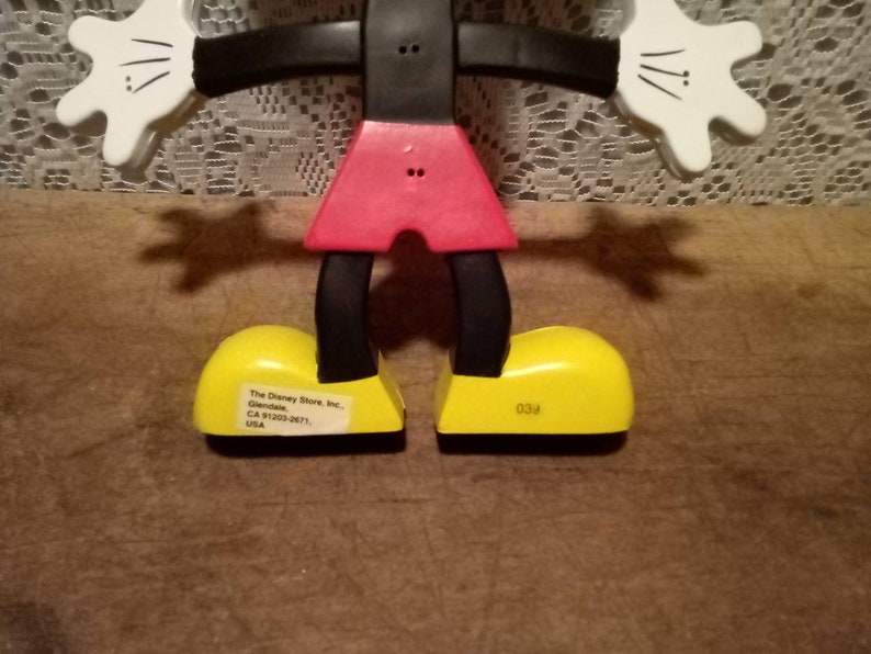 May include: A black and red articulated Mickey Mouse figurine with yellow shoes. The shoes have text on the bottom that reads "The Disney Store, Inc. Glendale, CA 91203-2671, USA" and "039".