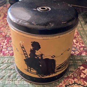 May include: Vintage yellow and black spool cotton tin with a black silhouette of a woman knitting. The tin has a black lid and a cylindrical shape. The text "Spool Cotton For Hand & Machine" is visible in the background.