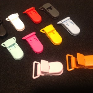 May include: A collection of colorful plastic pacifier clips in various shades, including black, white, red, gray, yellow, blue, green, pink, and orange. The clips are arranged on a dark surface.