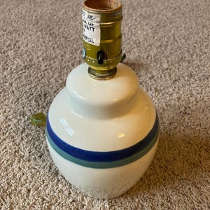 May include: A ceramic table lamp with a white base and blue and green stripes. The lamp has a brass-colored socket and a green handle. The label on the socket reads "TO REPLACE 100 WATT LAMP(S)."