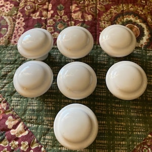 May include: Seven white ceramic cabinet knobs with a glossy finish are arranged on a patterned fabric background. The knobs are round with a slightly domed top and a flat base. The fabric has a floral design.