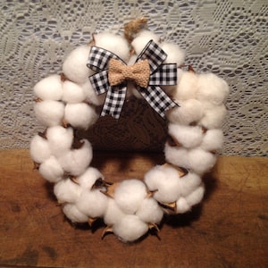 May include: A white cotton wreath with a black and white checkered bow and a burlap bow. The wreath is made of cotton bolls and is perfect for a farmhouse or rustic decor.