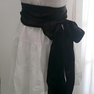May include: White dress with a square neckline, short puff sleeves, and a tiered skirt. A wide black sash is tied around the waist, creating a defined silhouette. The dress is displayed on a mannequin.