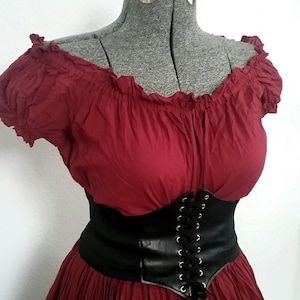 May include: A red off-the-shoulder peasant top with a black leather corset belt. The top has a ruffled neckline and short sleeves. The corset belt has a lace-up front.