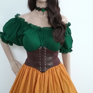 May include: A green off-the-shoulder peasant top with a brown leather corset and a long, tiered orange skirt. The model is wearing a flower crown and a green gemstone necklace.