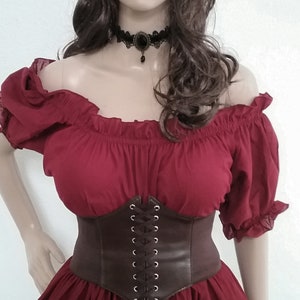 May include: A red off-the-shoulder peasant top with a brown leather corset belt. The top has a gathered neckline and short sleeves. The corset belt has a lace-up front with silver eyelets.