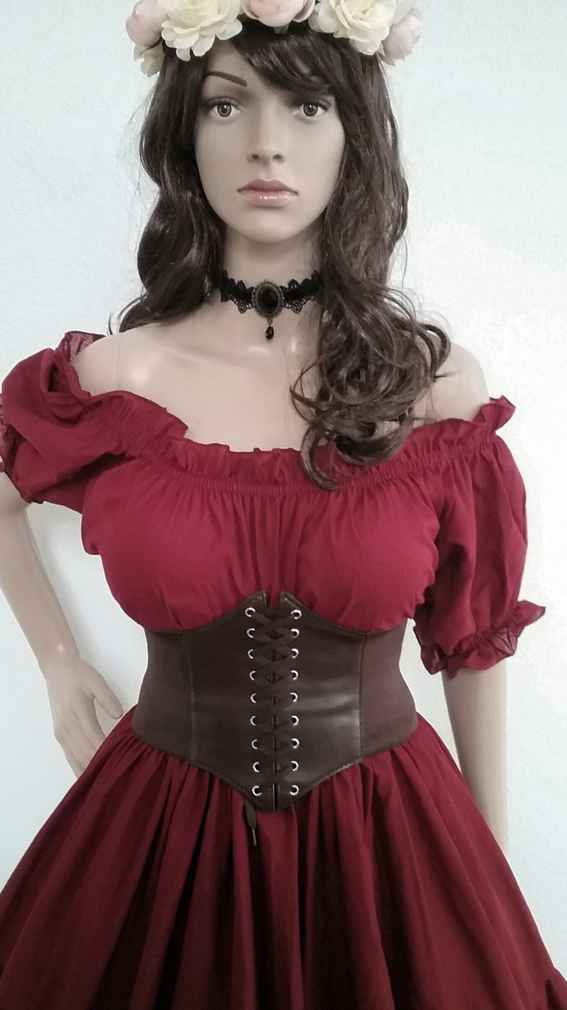 May include: A red off-the-shoulder peasant top with a brown leather corset belt. The top has a gathered neckline and short sleeves. The corset belt has a lace-up front with silver eyelets.