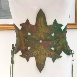 May include: A wide corset-style belt featuring a series of overlapping leaf-shaped panels in shades of green, brown, and hints of red. The panels are connected by small metallic studs, and the sides are secured with thin green ribbon lacing.