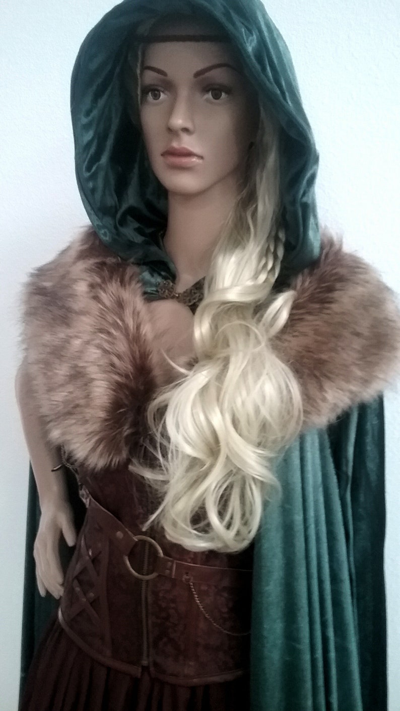 Puede incluir: Una mujer con una capa de terciopelo verde con capucha y cuello de piel marr&oacute;n. Lleva un cors&eacute; de cuero marr&oacute;n con una hebilla de metal. Su cabello rubio est&aacute; trenzado.