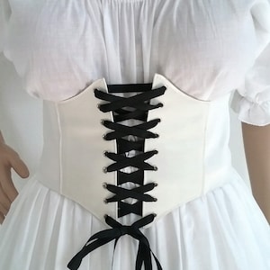 K&ouml;nnte beinhalten: Ein wei&szlig;es Korsett mit schwarzer Schn&uuml;rung &uuml;ber einem wei&szlig;en Kleid. Das Korsett hat ein strukturiertes Design mit spitzer Taille und wird mit schwarzem Band geschn&uuml;rt. Das Kleid hat einen schulterfreien Ausschnitt und einen flie&szlig;enden Rock.