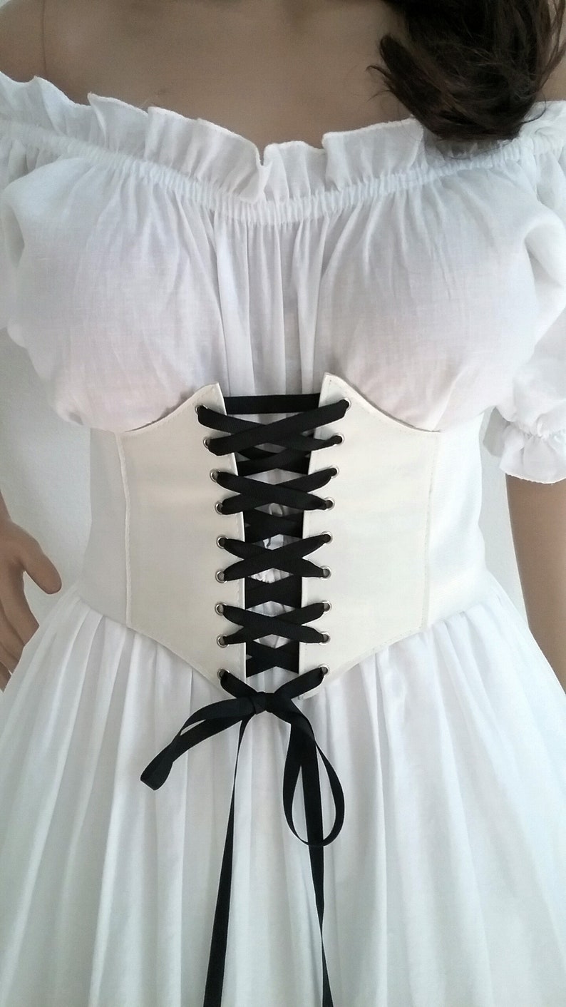 K&ouml;nnte beinhalten: Ein wei&szlig;es Korsett mit schwarzer Schn&uuml;rung &uuml;ber einem wei&szlig;en Kleid. Das Korsett hat ein strukturiertes Design mit spitzer Taille und wird mit schwarzem Band geschn&uuml;rt. Das Kleid hat einen schulterfreien Ausschnitt und einen flie&szlig;enden Rock.