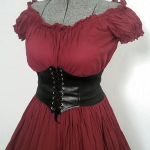 May include: A maroon off-the-shoulder dress with a black leather corset belt. The dress has a ruffled neckline and a tiered skirt.