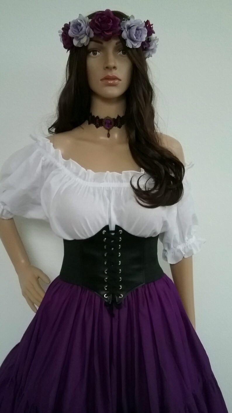 May include: A woman wearing a white peasant top with a black corset and a purple skirt. She is wearing a flower crown with purple and white flowers.