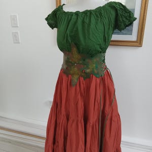 May include: A two-piece outfit consisting of a dark green off-the-shoulder blouse with short puffed sleeves and a long, tiered rust-colored skirt. A wide, leaf-shaped brown leather corset belt with green accents and side lacing cinches the waist.