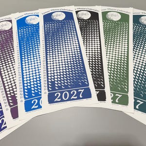 May include: A collection of seven vertical 2027 Lunar Phase Calendars arranged in a fan shape. Each paper calendar features a unique colour background, including blue, purple, teal, black, green, and olive, with white moon phase icons and numbers.