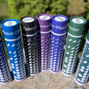 May include: A collection of cylindrical calendars for 2026, each with a different color scheme. The calendars are blue, black, purple, green, and teal, with white crescent moon patterns. The text "Lunar Phase Calendar" is visible on some.