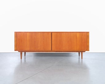 May include: A mid-century modern teak wood sideboard with two doors and tapered legs. The sideboard has a light wood finish and is in good condition.