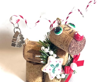 May include: A handmade Christmas ornament crafted from wine corks, featuring a reindeer design. The reindeer has googly eyes, a red nose, and a star-shaped decoration. Red and white twine serves as antlers, and a small tree charm hangs from the ornament.