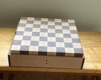 May include: A wooden chessboard with a checkerboard pattern in light and dark wood. The board is closed and sitting on a wooden table.